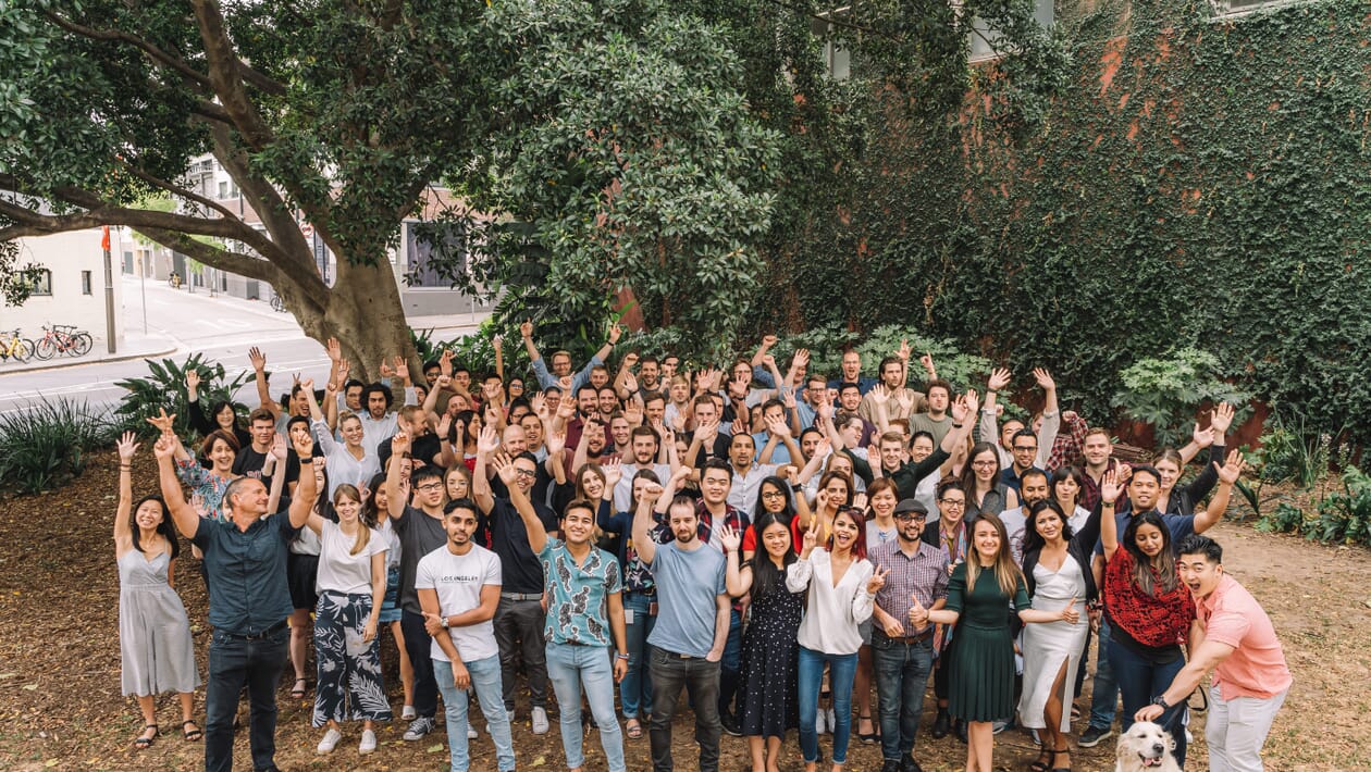 A group photo of all the people who work at the Sydney office.. There are around 100 people in the photo and a dog.